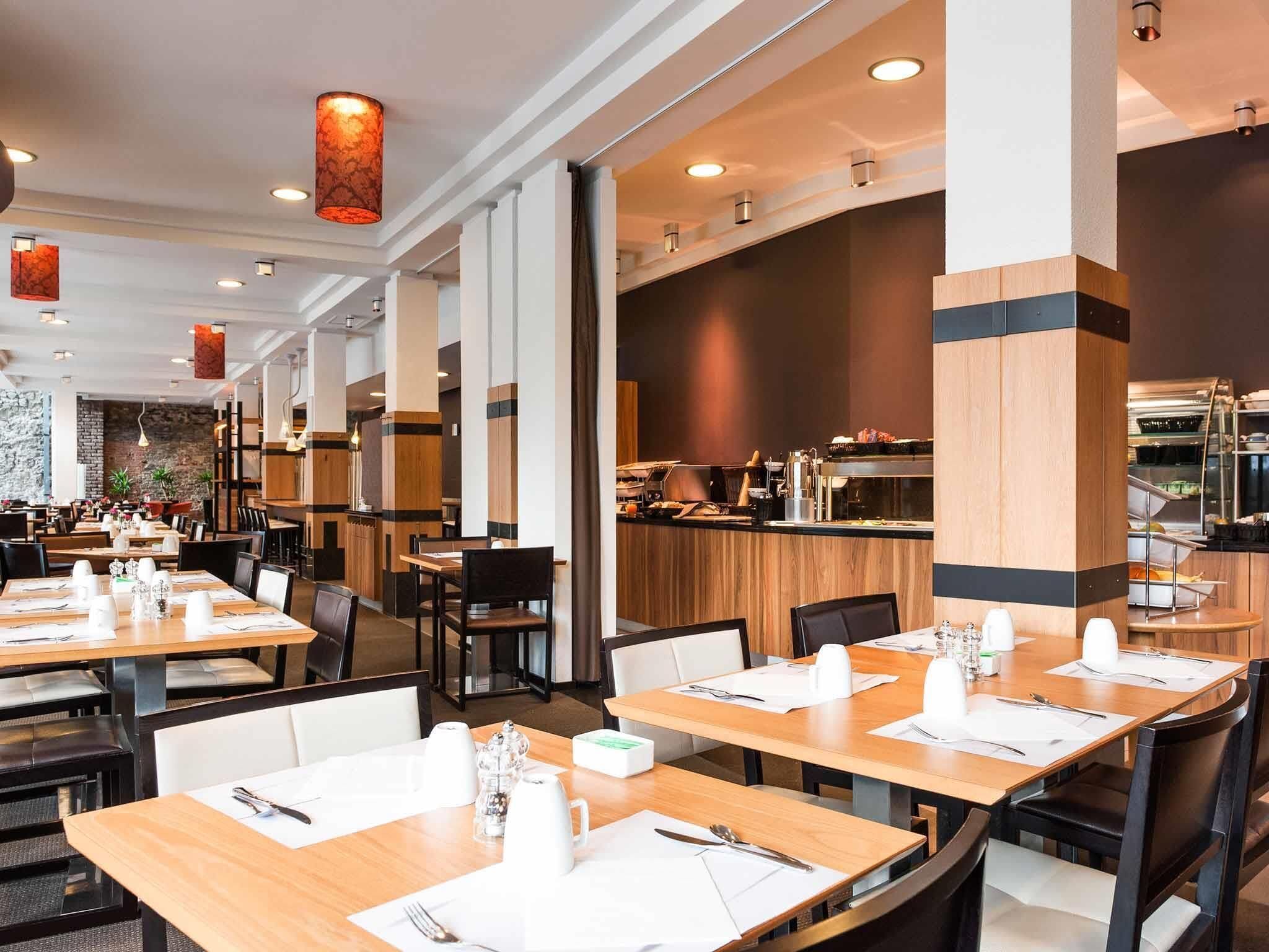 A modern restaurant with wooden tables and chairs, a kitchen area in the background, and pendant lighting.