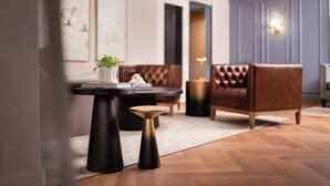 Un salon moderne avec un canapé en cuir marron, une table basse noire et un petit tabouret. On trouve une plante en pot sur la table basse et une lampe sur une table d’appoint.