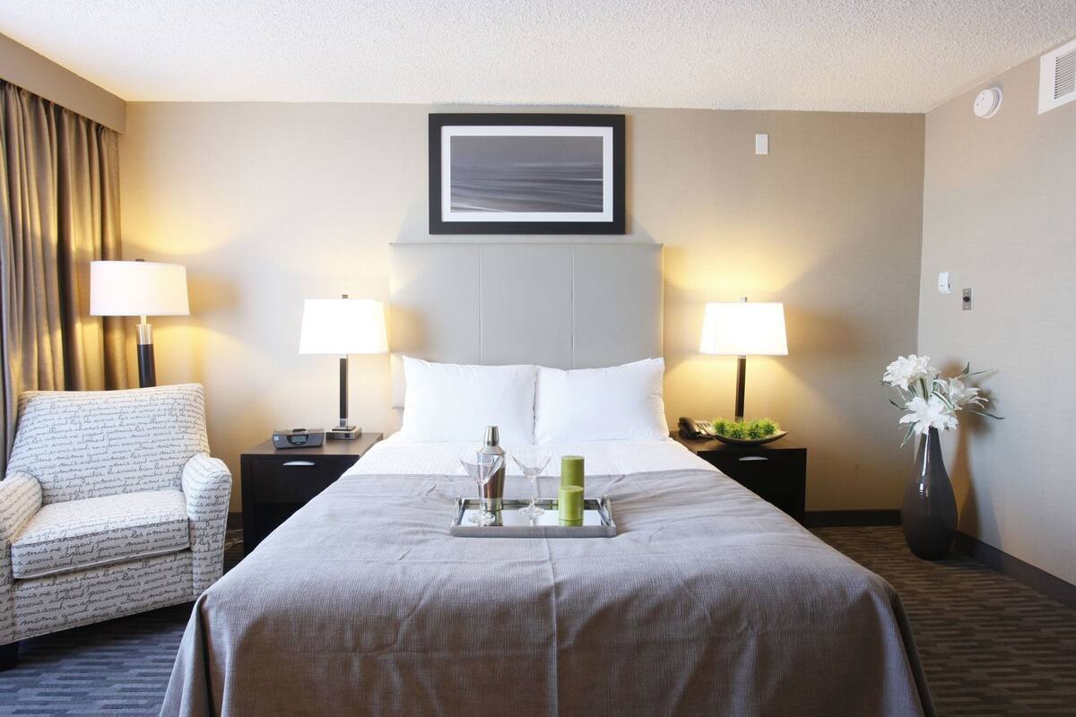 A hotel room with a bed, two bedside tables, a chair, a framed picture on the wall, and a vase with flowers.