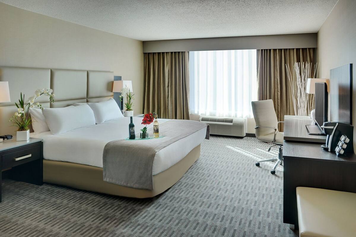 A hotel room with a bed, bedside tables, a desk, a chair, a TV, and a large window with curtains.