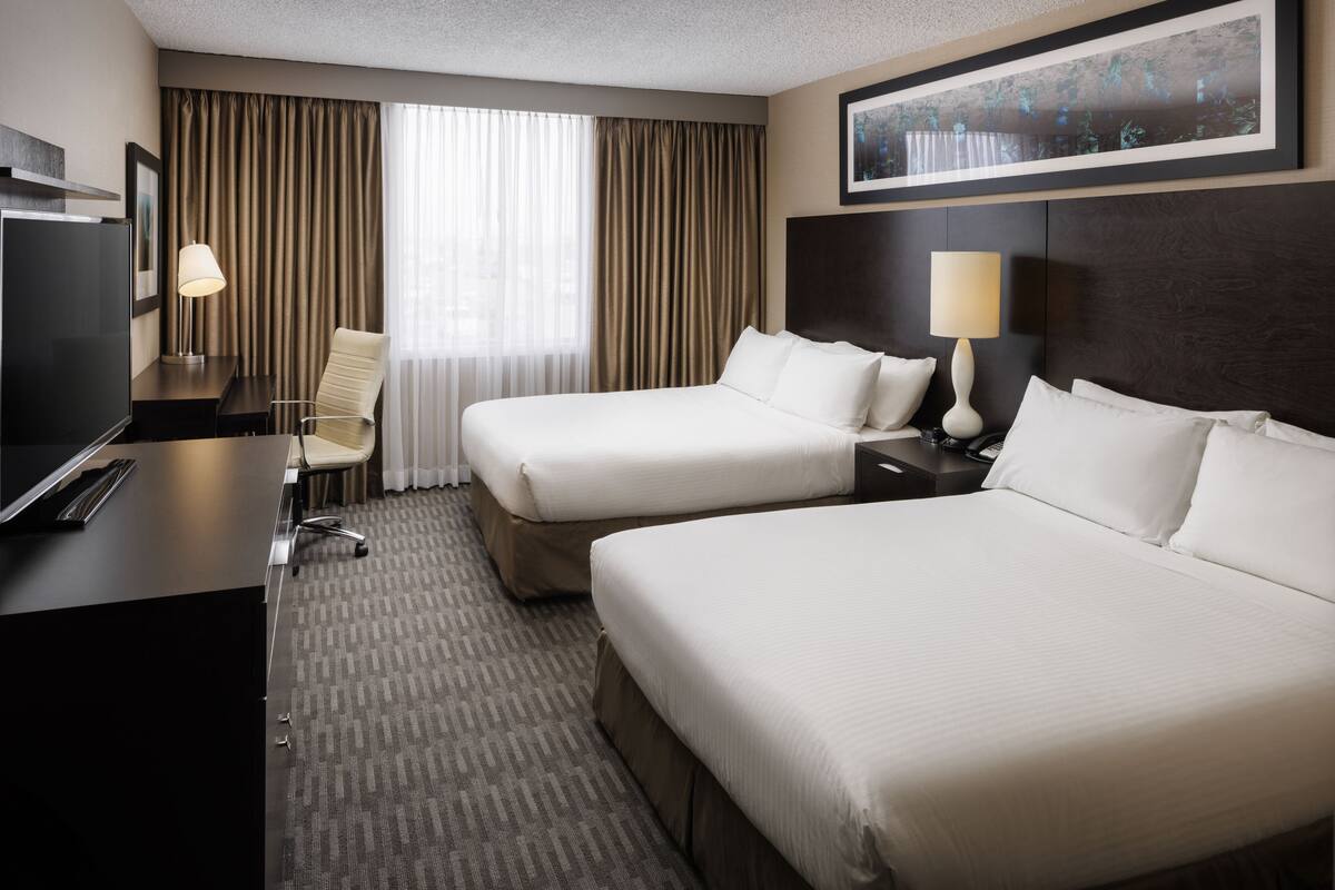 A hotel room with two beds, a desk, a chair, a TV, and a large window with curtains.