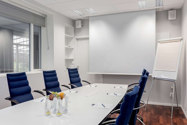 Un salón de conferencias con una mesa blanca, sillas azules, una pizarra y una ventana con persianas.