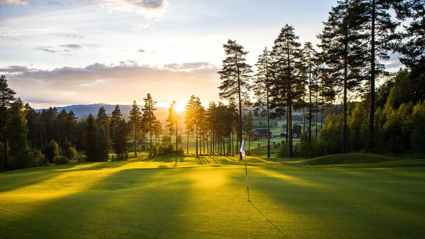 A golf course with a flagstick, surrounded by tall trees and a sunset in the background. - Best Western Plus Gyldenlove Hotell (Kongsberg)
