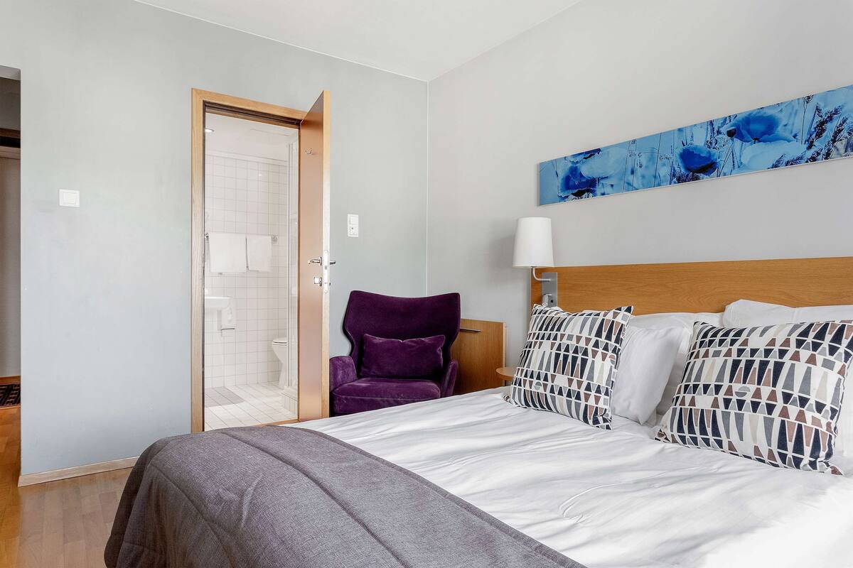 Une chambre à coucher avec un lit, un fauteuil violet, un tableau et une lampe blanche.