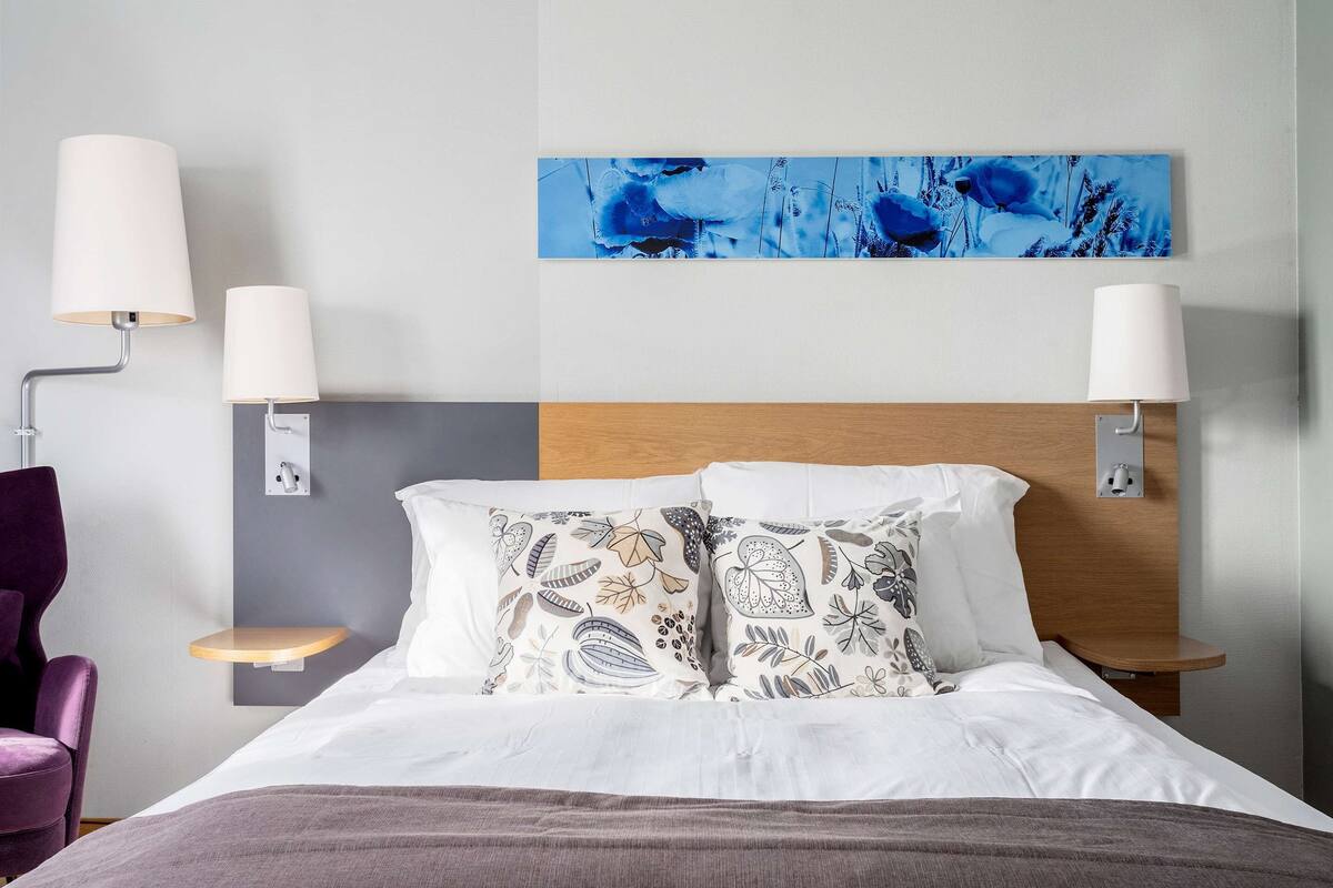 Une chambre d’hôtel comprenant un lit, deux tables de chevet, une chaise, une lampe fixée au mur et un tableau.
