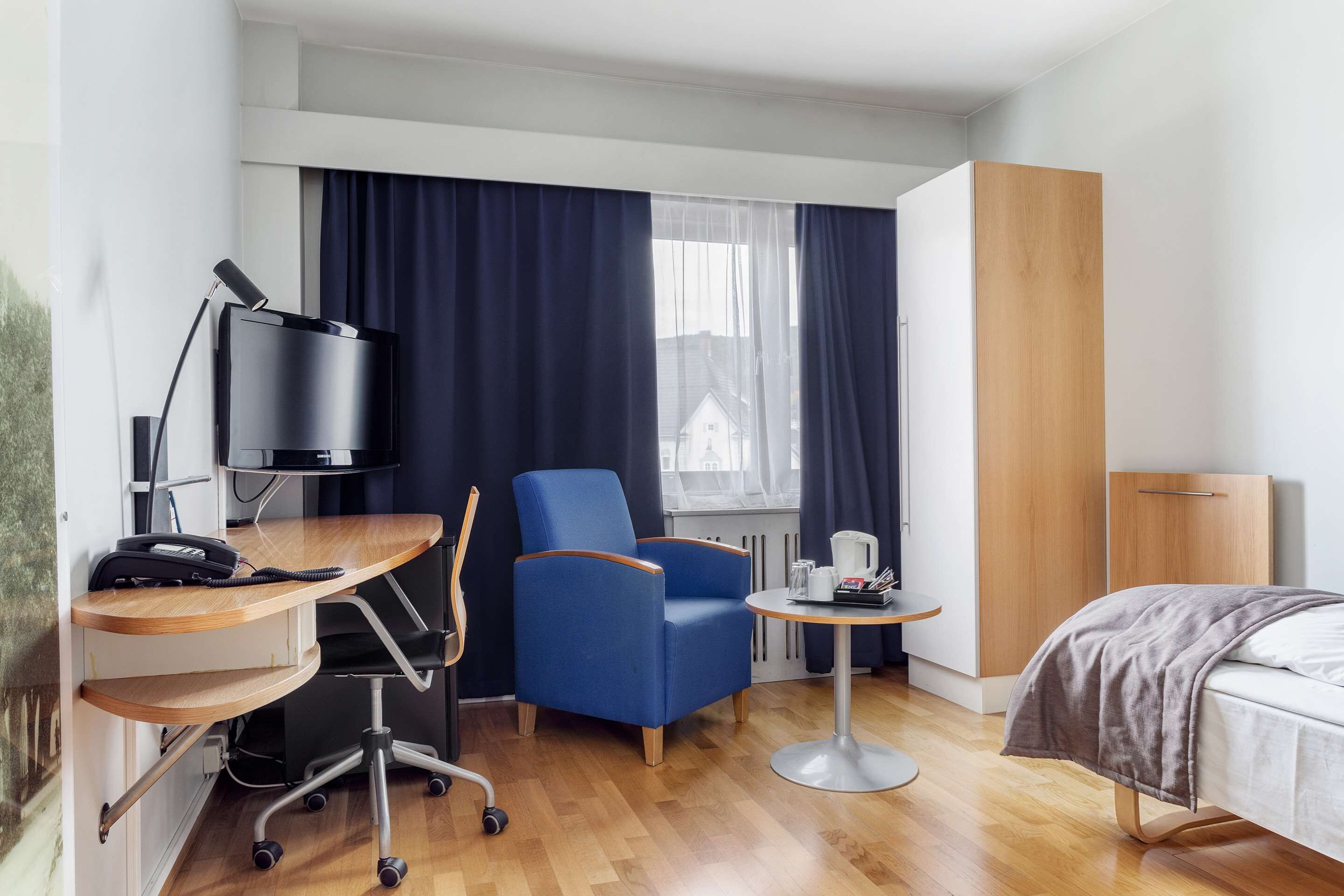 A hotel room with a desk, chair, bed, and a small table with a kettle.