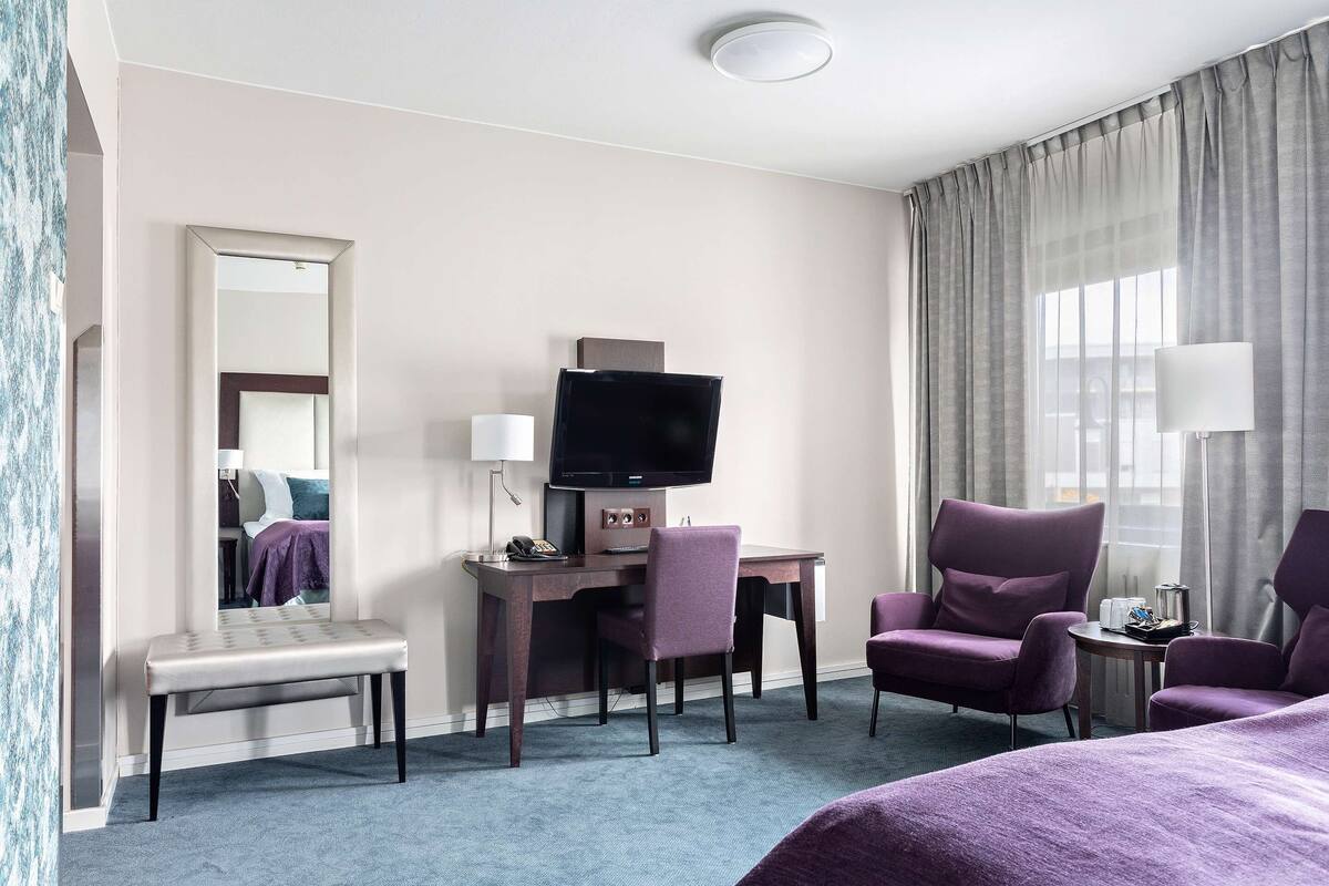 Une chambre d’hôtel équipée d’un bureau, d’une télévision, d’une chaise, d’un lit et d’une fenêtre avec des rideaux.