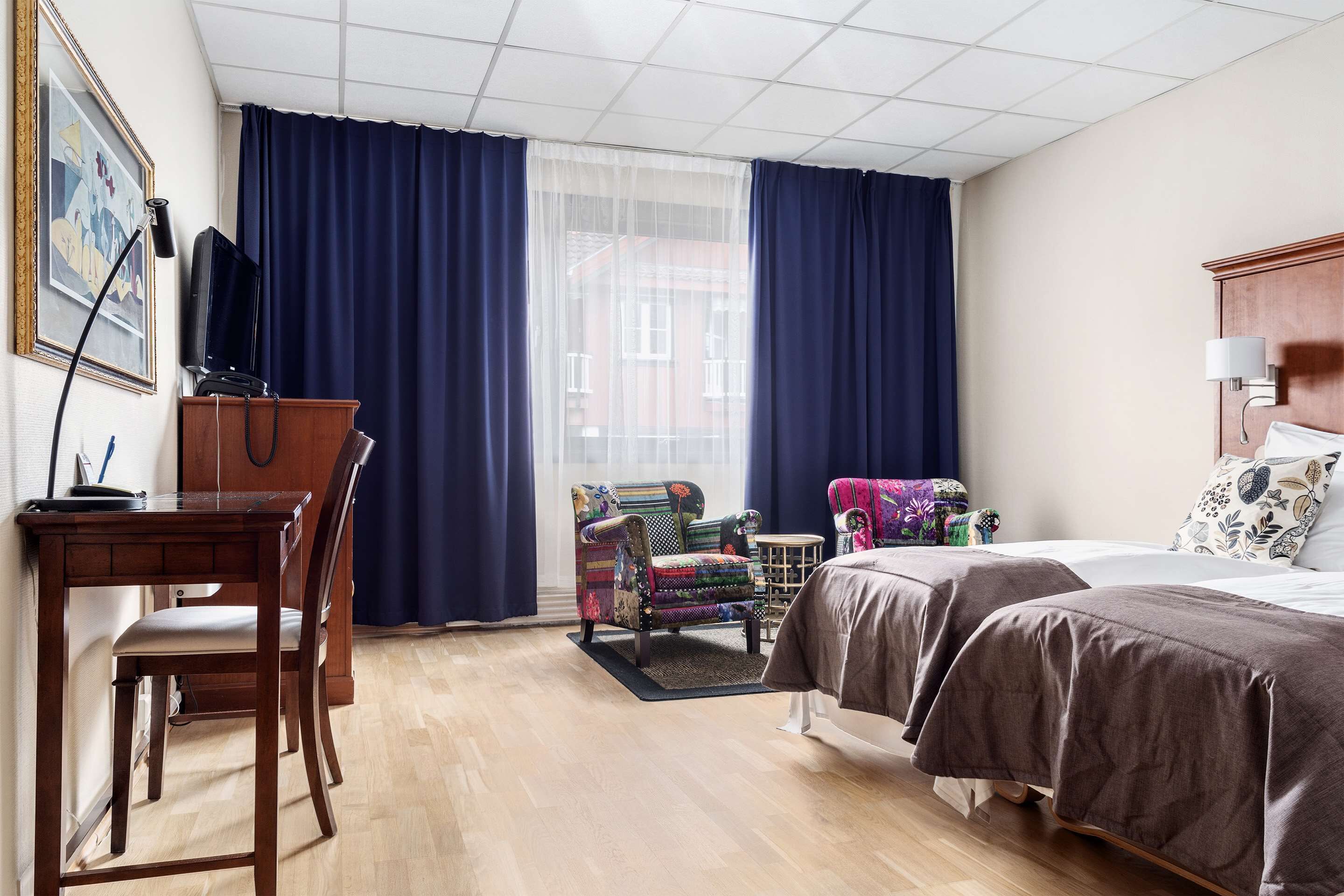 A hotel room with a wooden desk, a colorful armchair, a bed with two beds, and a large window with blue curtains.