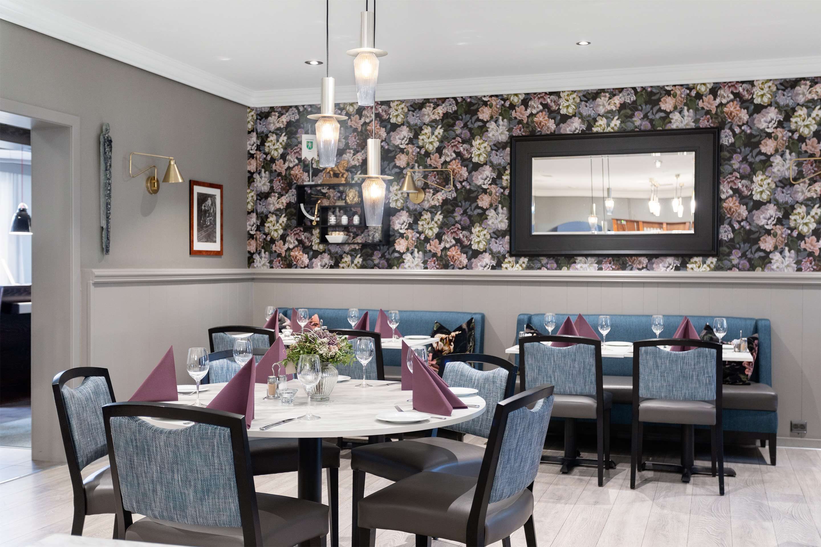 Un comedor con una mesa redonda dispuesta para cuatro personas, sillas tapizadas en azul y un fondo de papel tapiz floral.