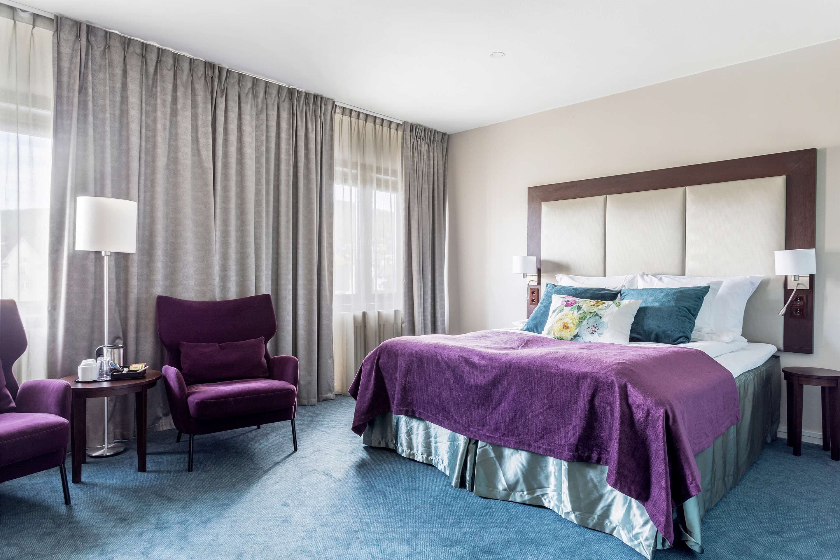 A hotel room with a large bed, a purple armchair, a small table with a lamp, and a large window with curtains.