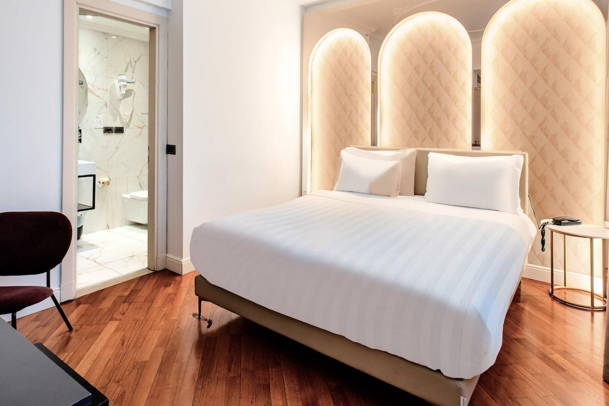 A hotel room with a large bed, a wooden floor, a modern chair, and a bathroom visible through an open door.