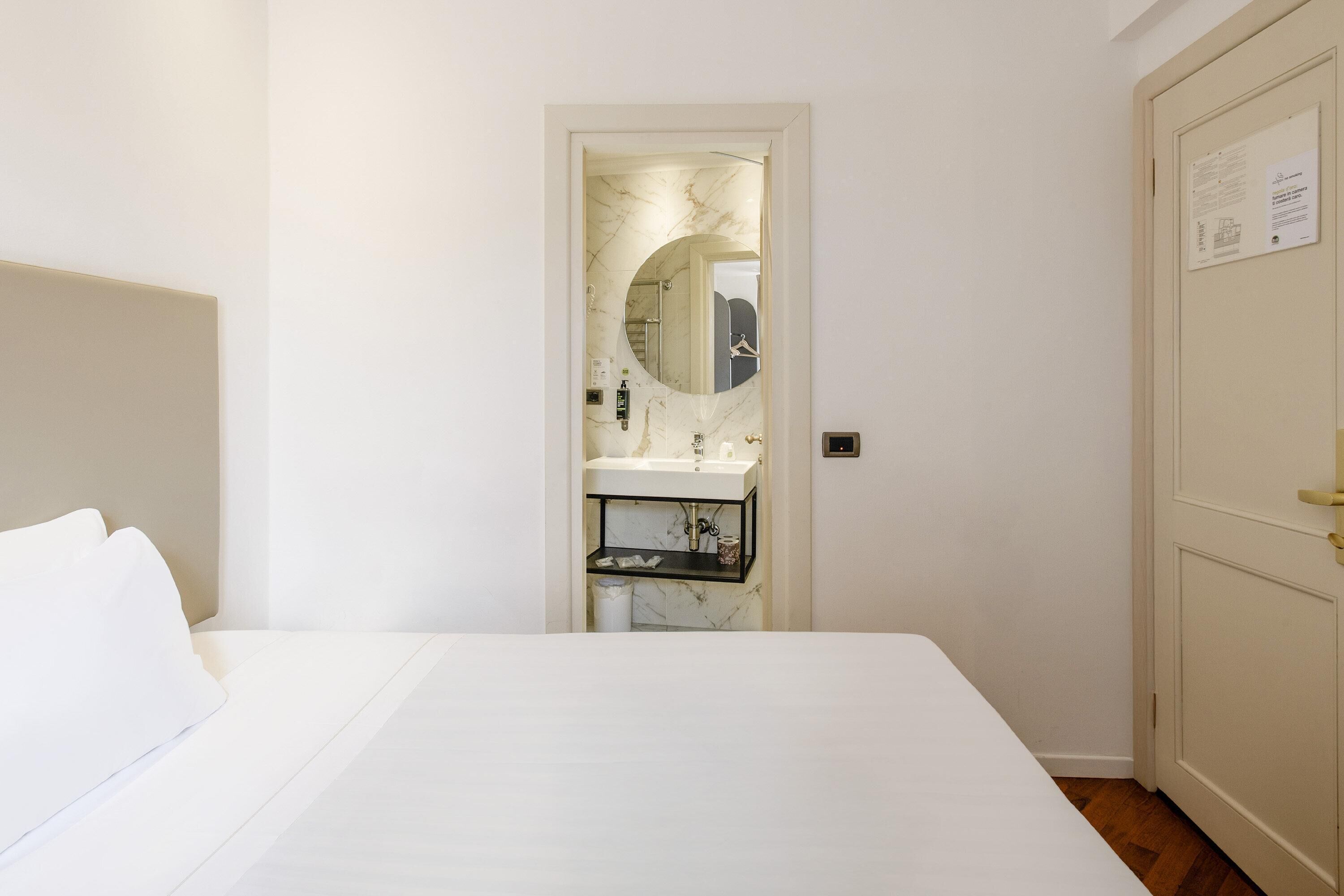Una habitación de hotel con cama, baño con lavamanos y espejo, y puerta con un cartel.