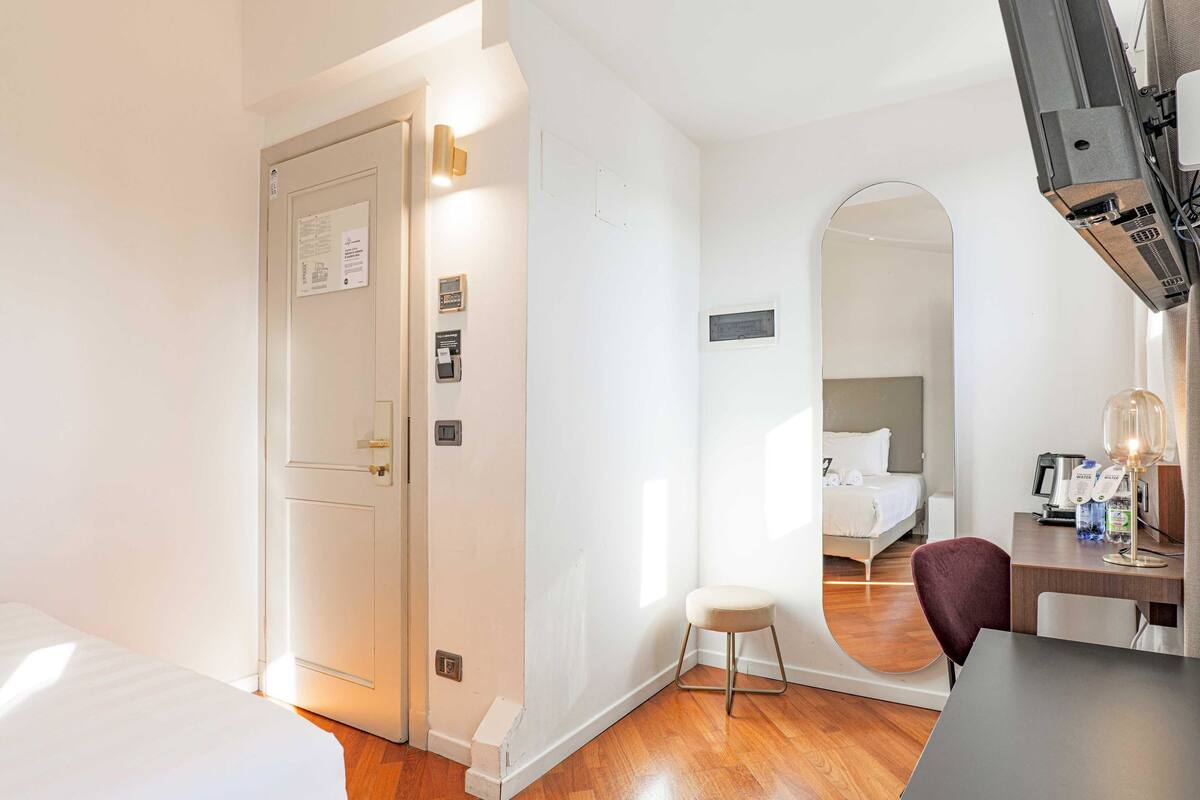 A hotel room with a wooden floor, a bed, a desk with a lamp, and a mirror.