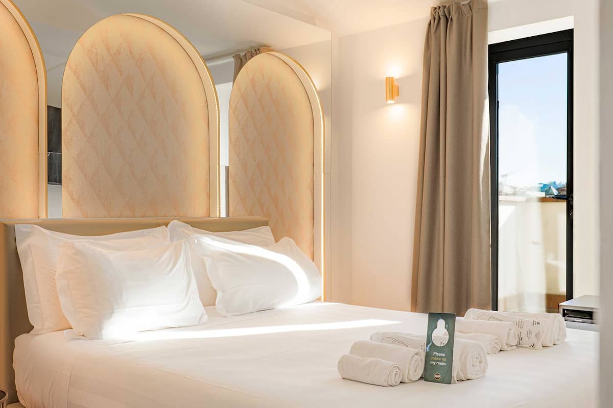 A hotel room with a large bed, a headboard, a window with curtains, and a set of towels on the bed.