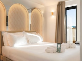 Quarto de hotel com uma cama grande, cabeceira, janela com cortinas e um conjunto de toalhas na cama.