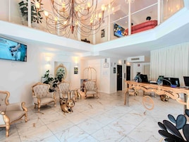 Um hall amplo com teto alto, um candelabro e uma televisão na parede. Há várias cadeiras e uma mesa com computadores.
