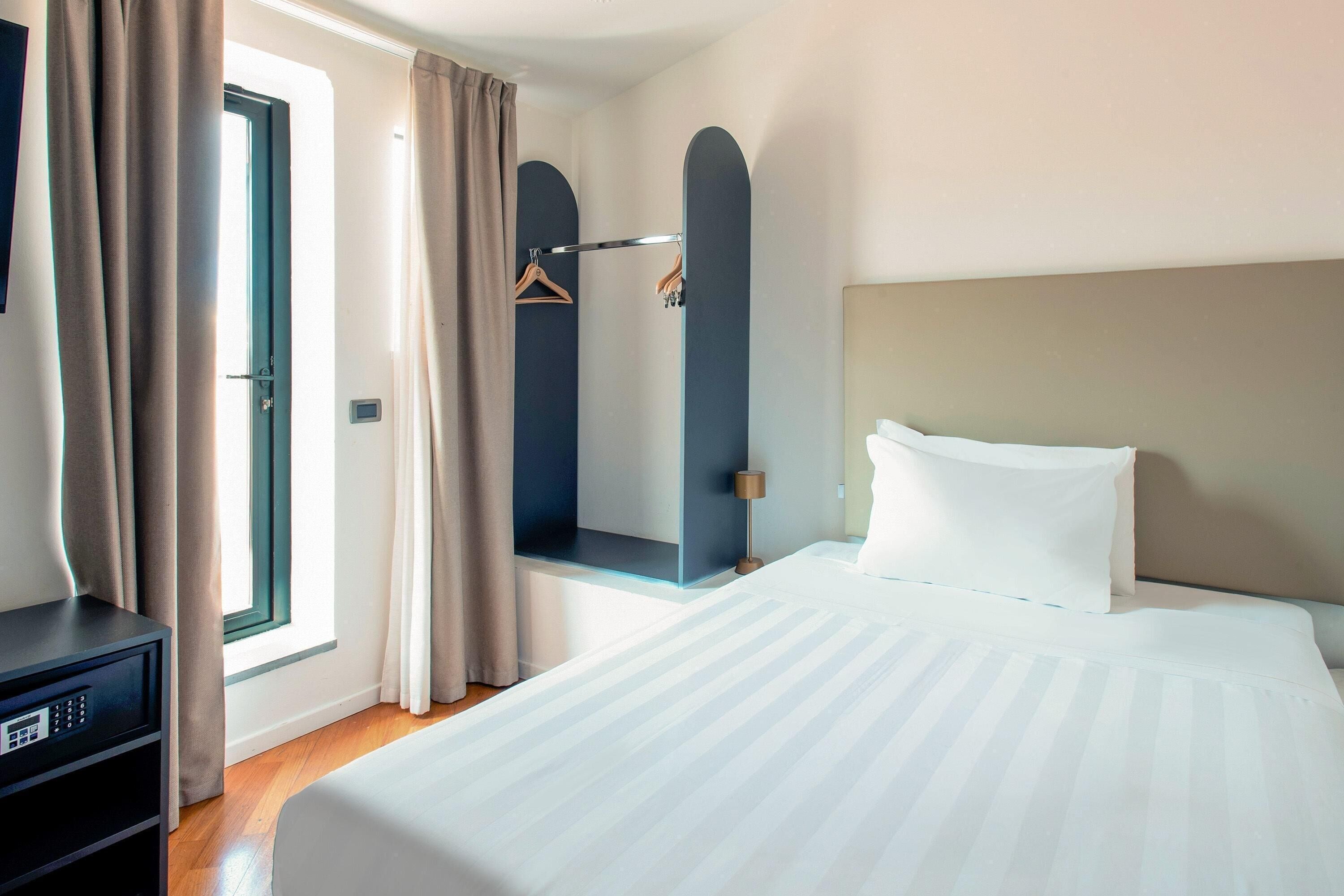 A hotel room with a bed, a desk, a hanging coat rack, and a window with curtains.