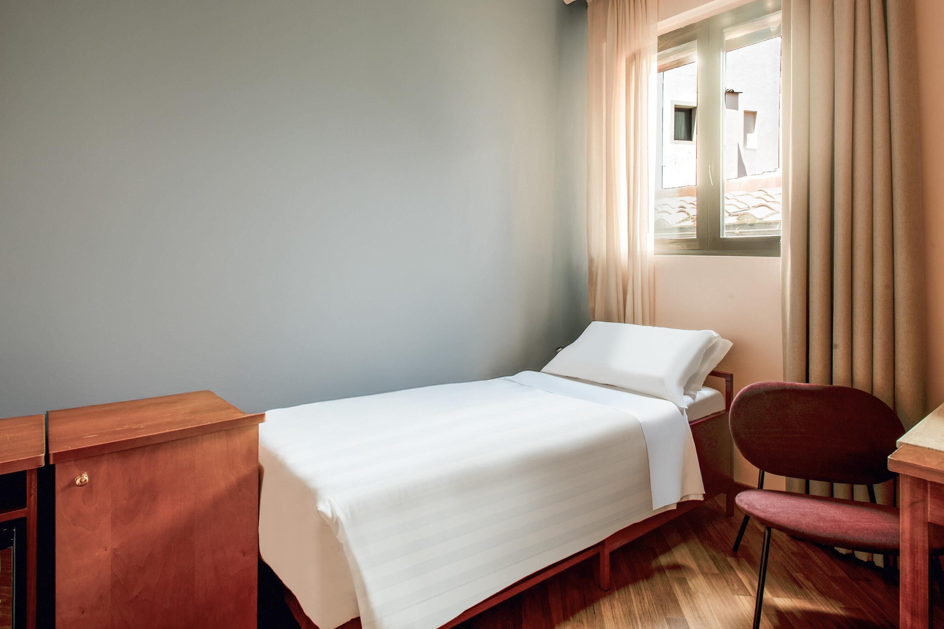 Un lit simple avec une couvre-lit blanche, une table de chevet en bois, une chaise et une fenêtre avec des rideaux.
