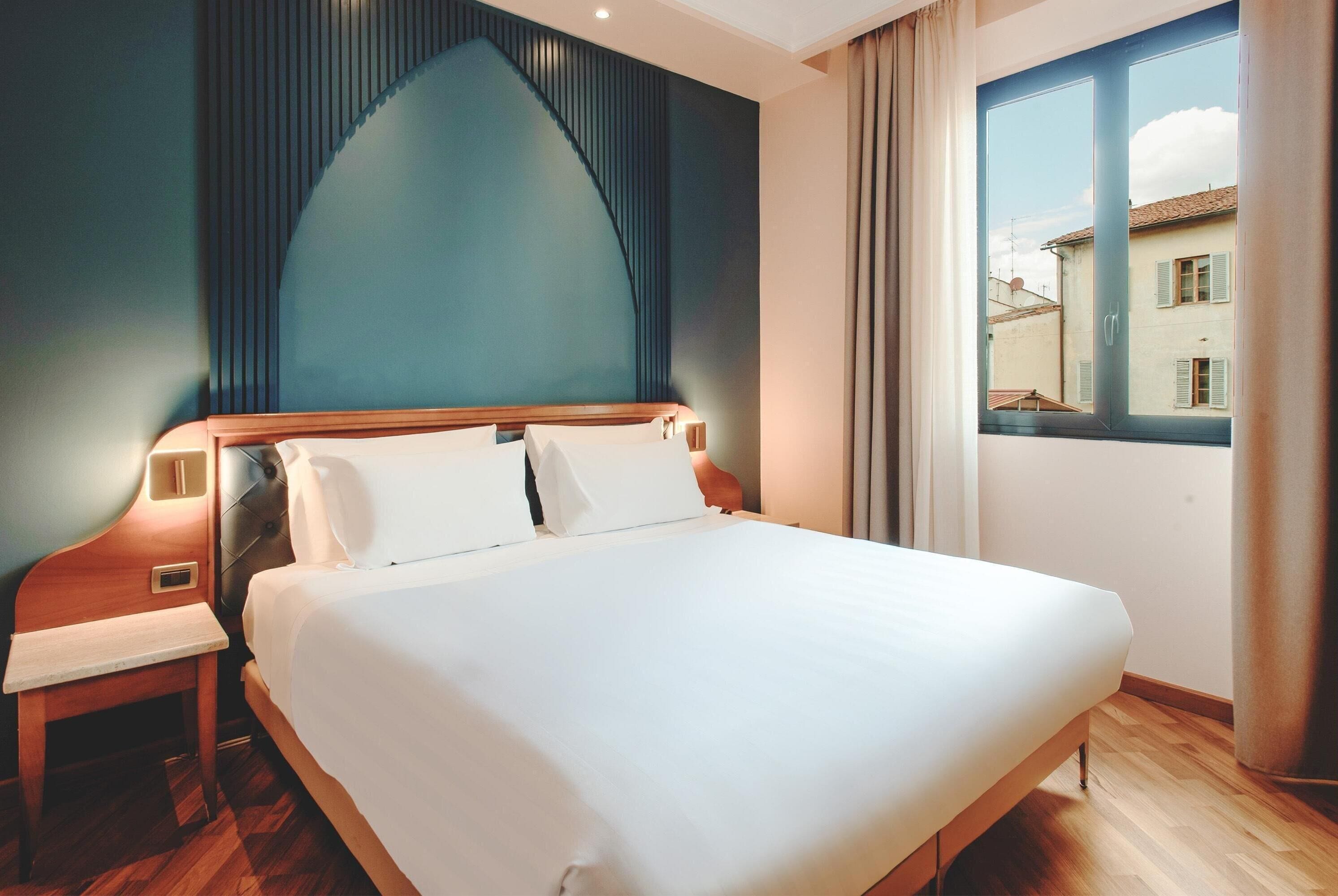 Quarto de hotel com uma cama grande, uma mesinha de cabeceira de madeira e vista para prédios pela janela.