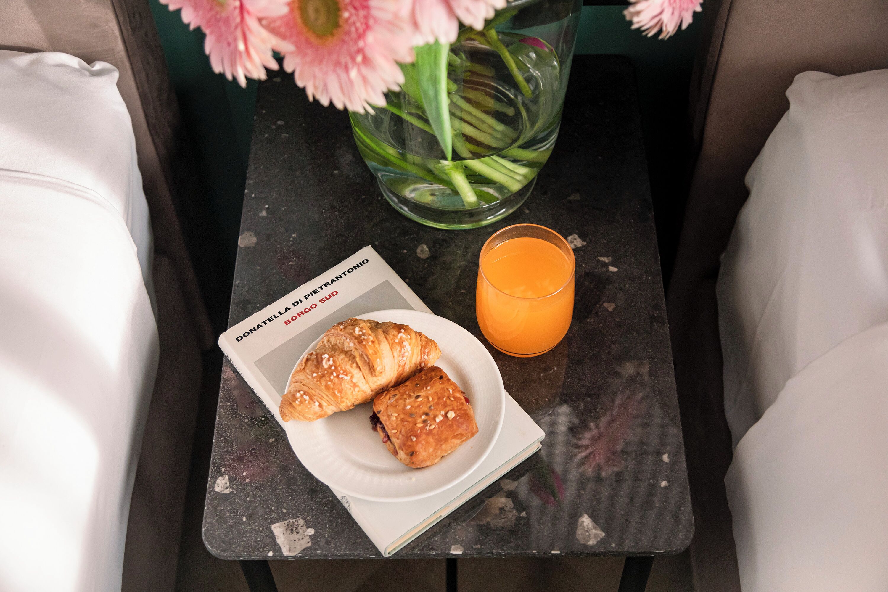 A breakfast setup with croissants on a plate, a glass of orange juice, and a book titled "Donatella P. Borgo Santonio" on a small table.
