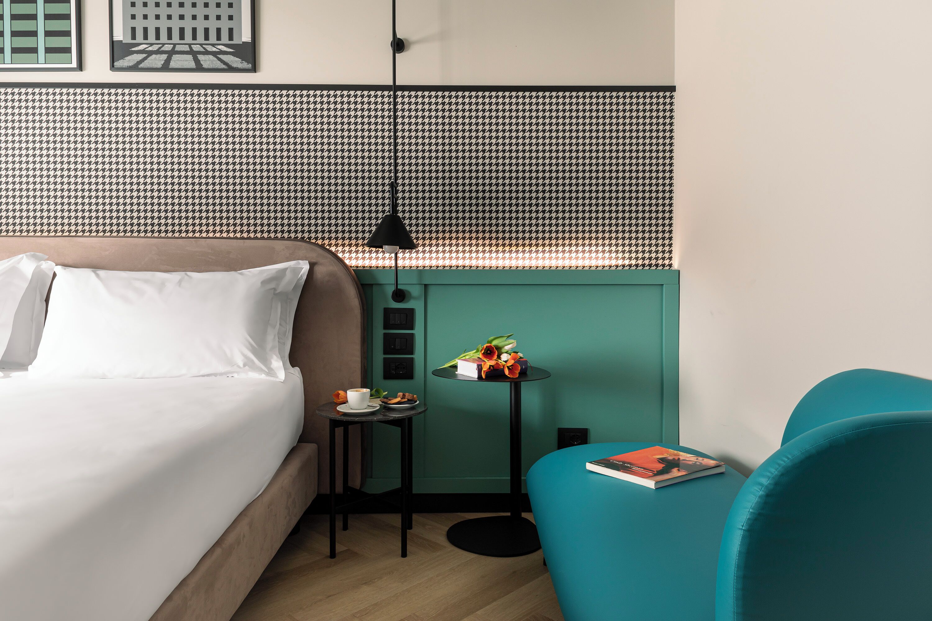 Habitación de hotel ordenada con cama, una mesita con una taza y un libro, una silla color turquesa y una pared con estampado de piedritas.