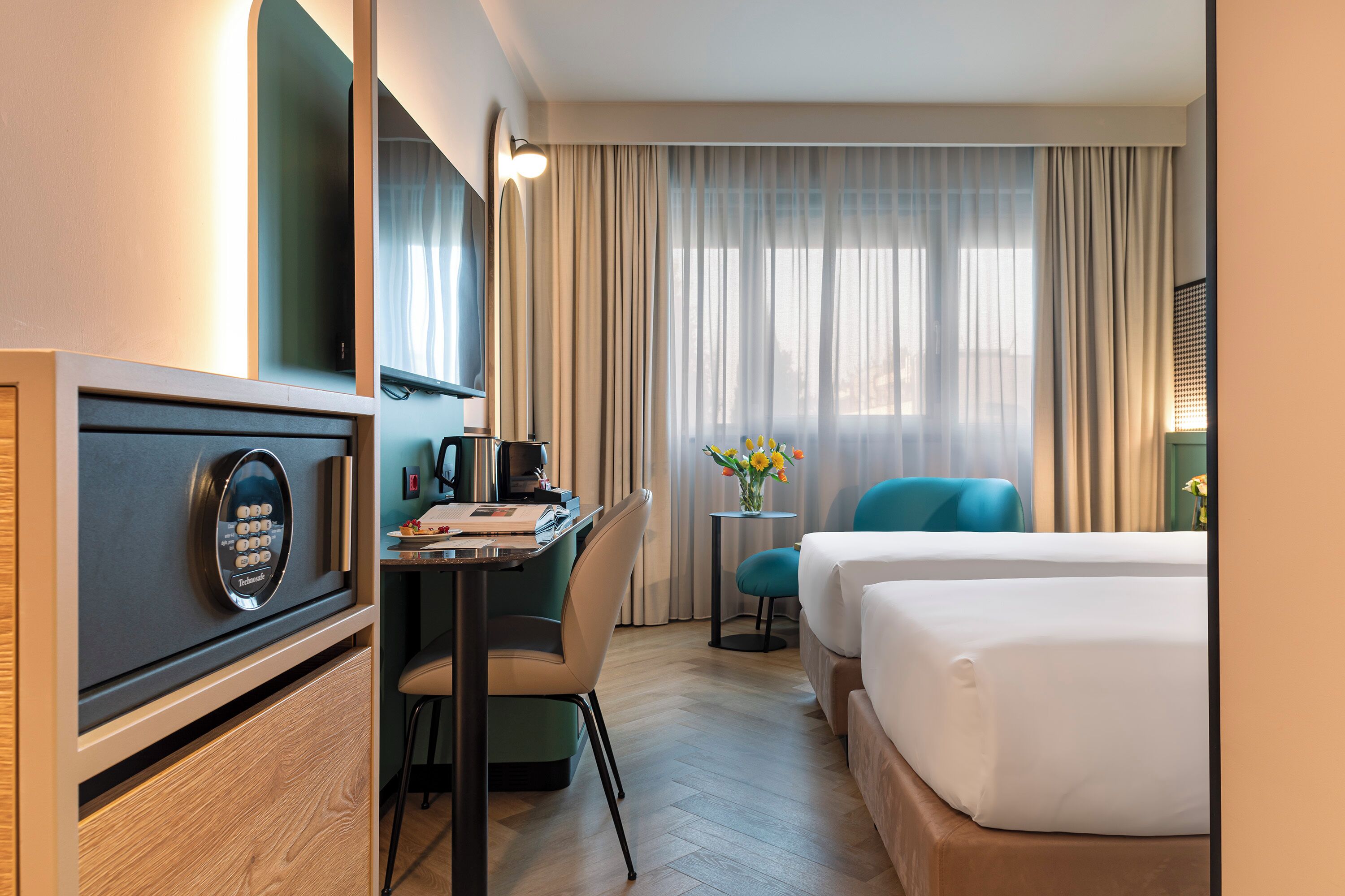 A hotel room with a bed, a desk with a computer, a chair, a small table with a vase, and a window with curtains.