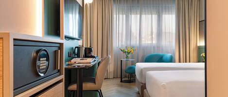 A hotel room with a bed, a desk with a computer, a chair, a small table with a vase, and a window with curtains.