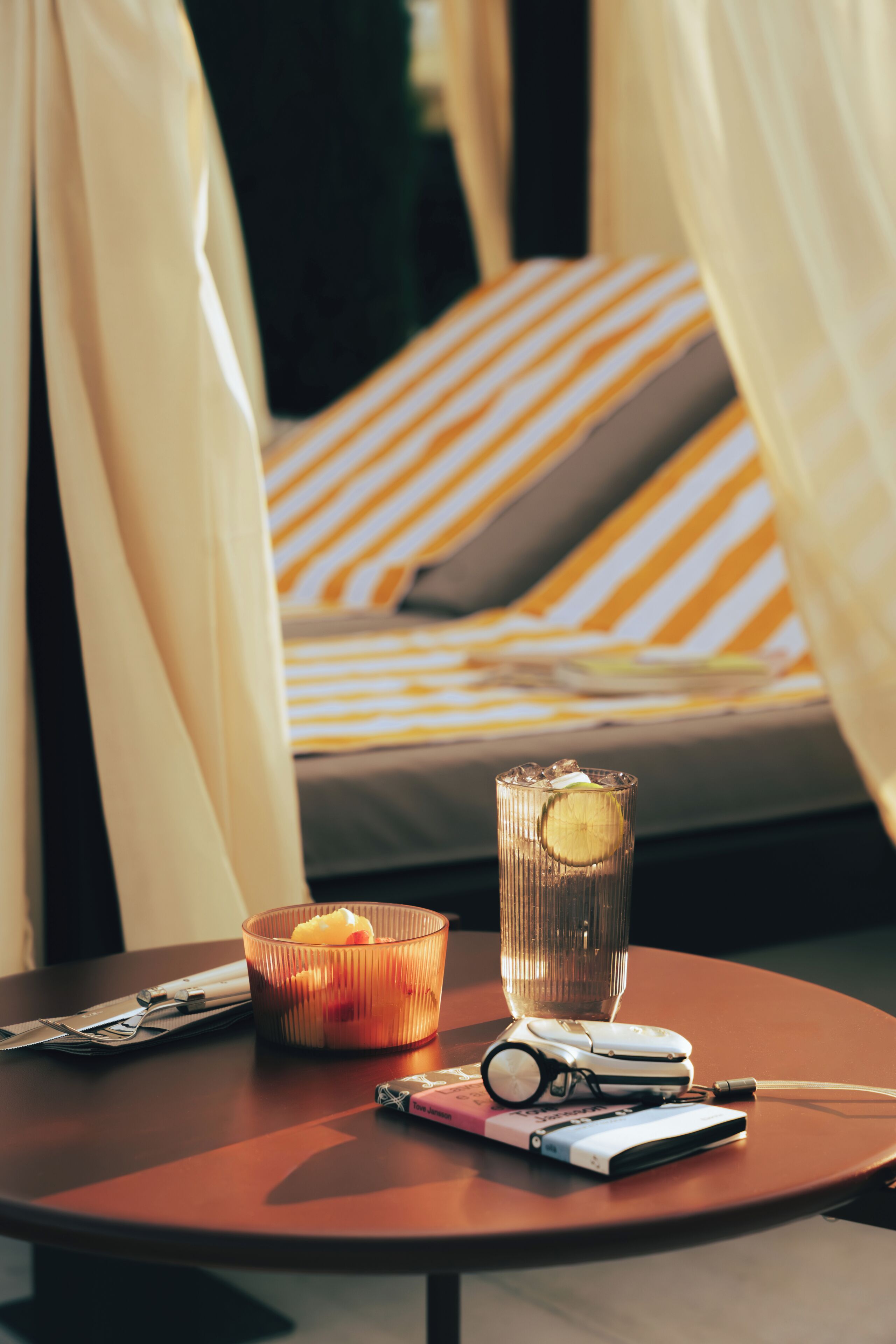 A bed with striped bedding, a glass of water with a slice of lime, a candle, and a pair of glasses on a table.