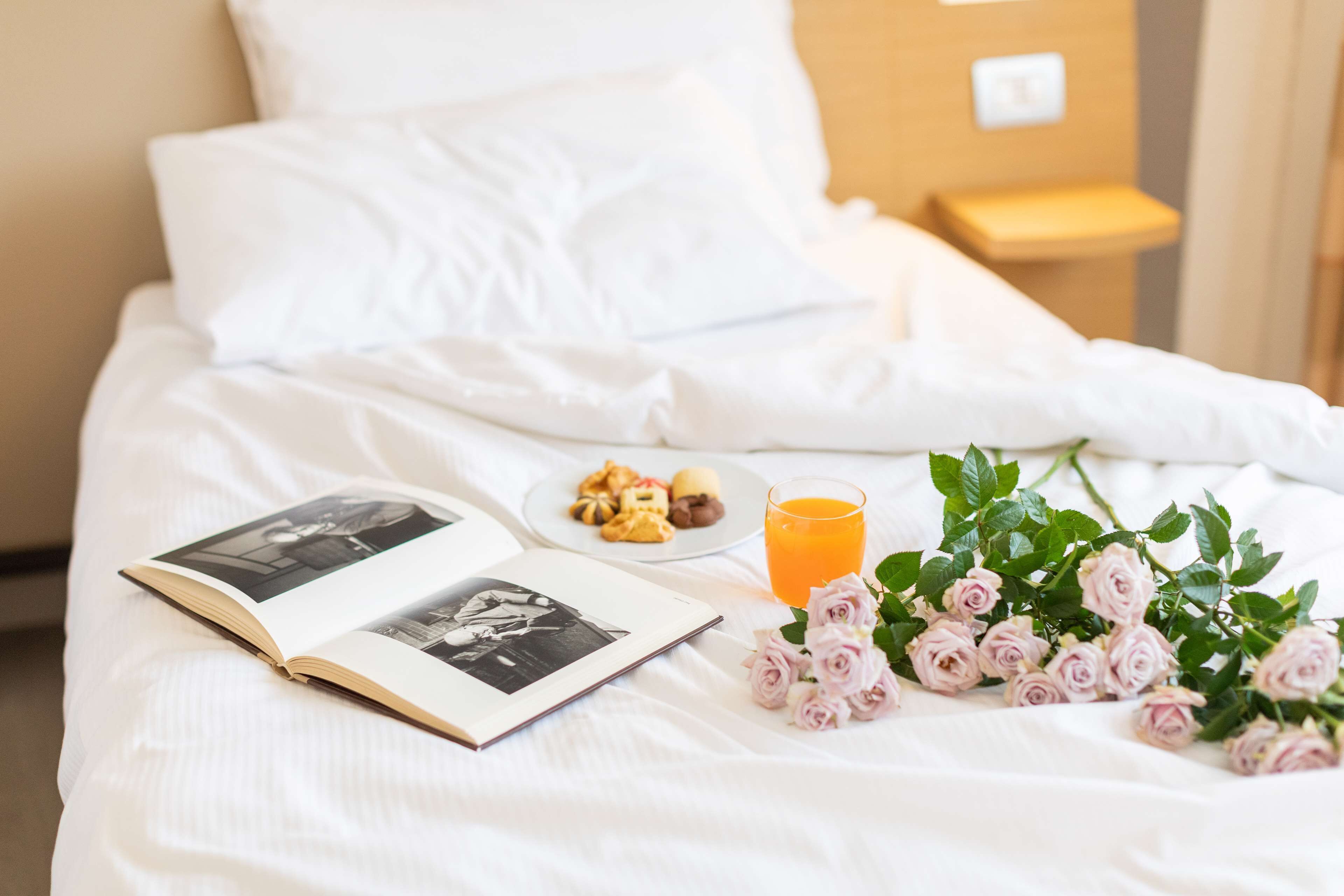 A neatly made bed with white linens, a book, a plate of pastries, a glass of orange juice, and a bouquet of pink roses.