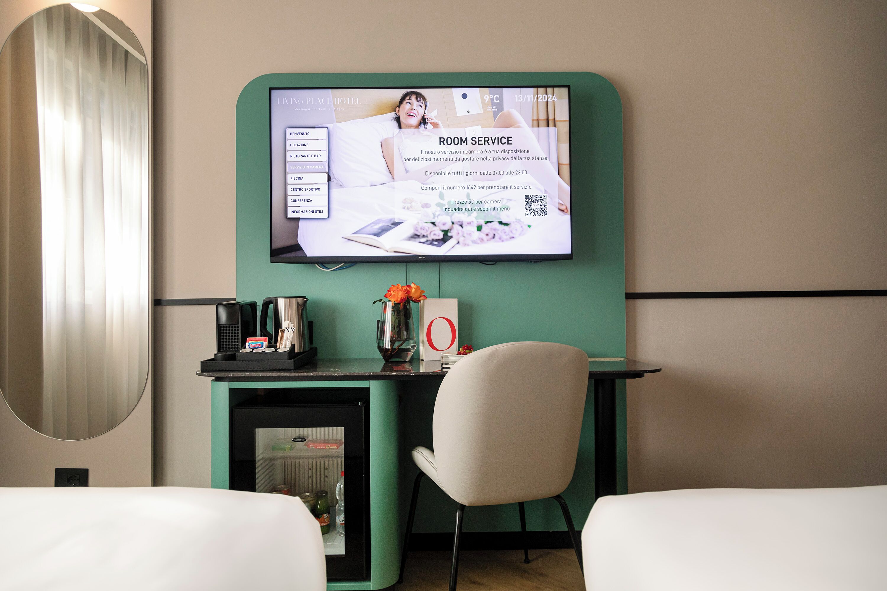A hotel room with a TV displaying room service information, a desk with a coffee machine, a vase with flowers, and a chair.