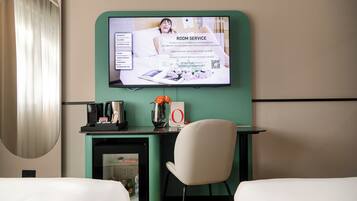 A hotel room with a TV displaying room service information, a desk with a coffee machine, a vase with flowers, and a chair.