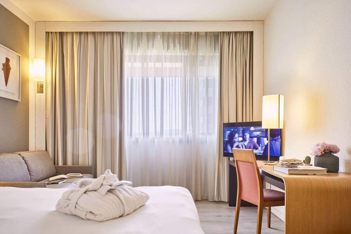 Ein Hotelzimmer mit einem Bett, einem Schreibtisch mit Stuhl, einem Fernseher und einem Fenster mit Vorhängen.