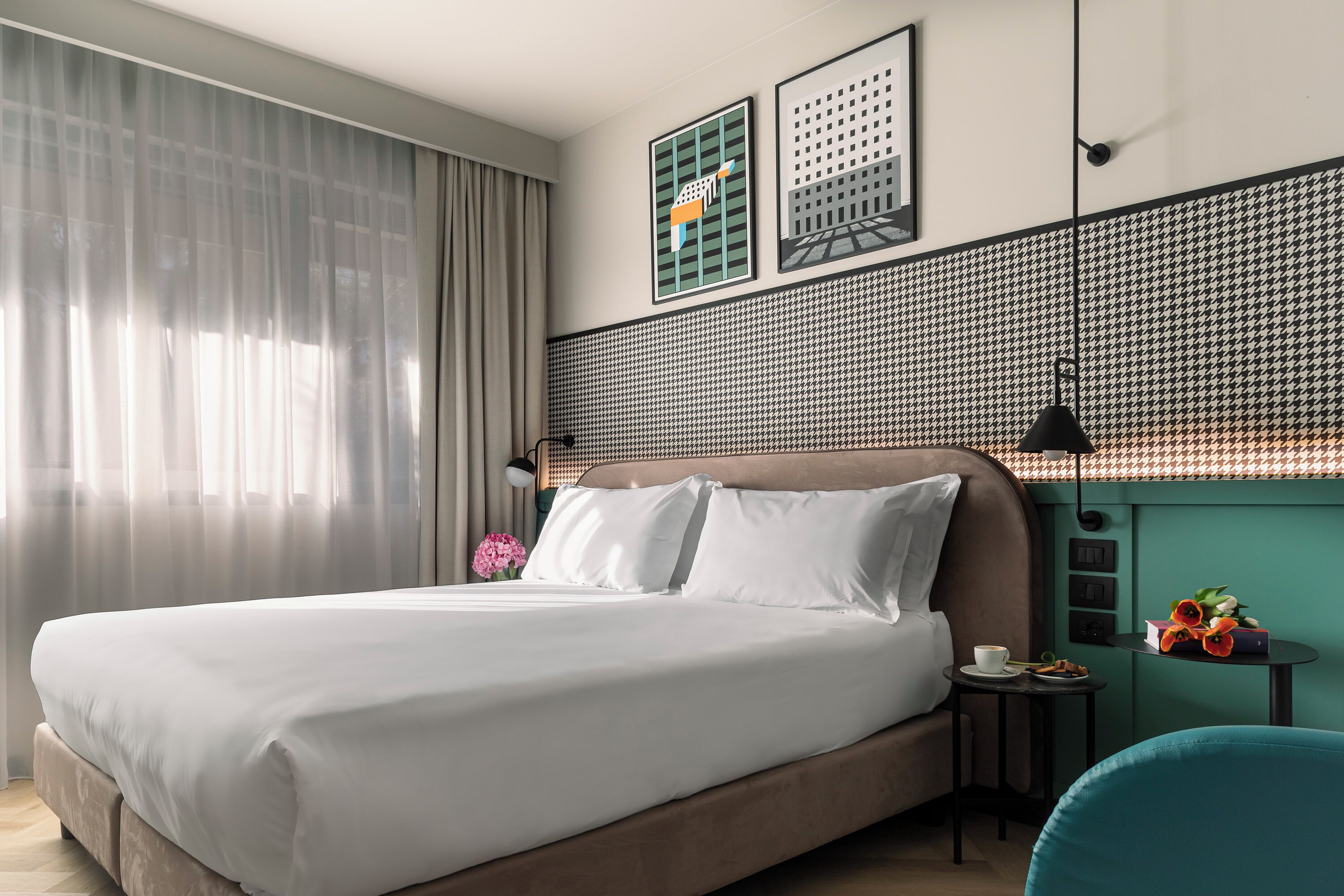 Une chambre d’hôtel moderne avec un grand lit, une petite table sur laquelle se trouvent une tasse et des fleurs, et une vue sur la ville par la fenêtre.