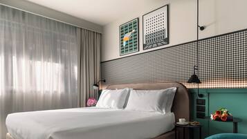 A modern hotel room with a large bed, a small table with a cup and flowers, and a view of the city through the window.