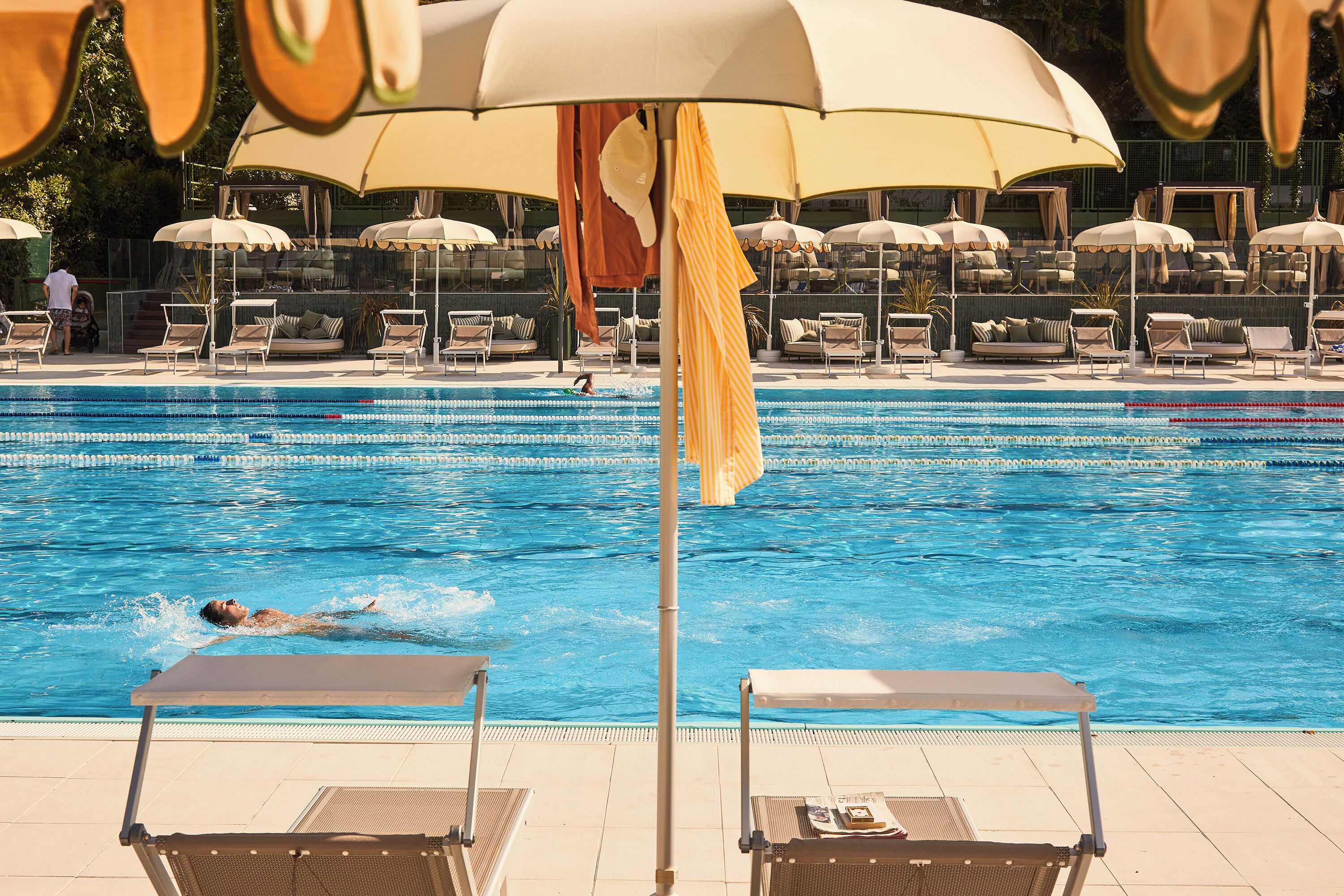 Piscina con sillones de descanso y sombrillas, una persona nadando y una zona de terraza con baldosas.