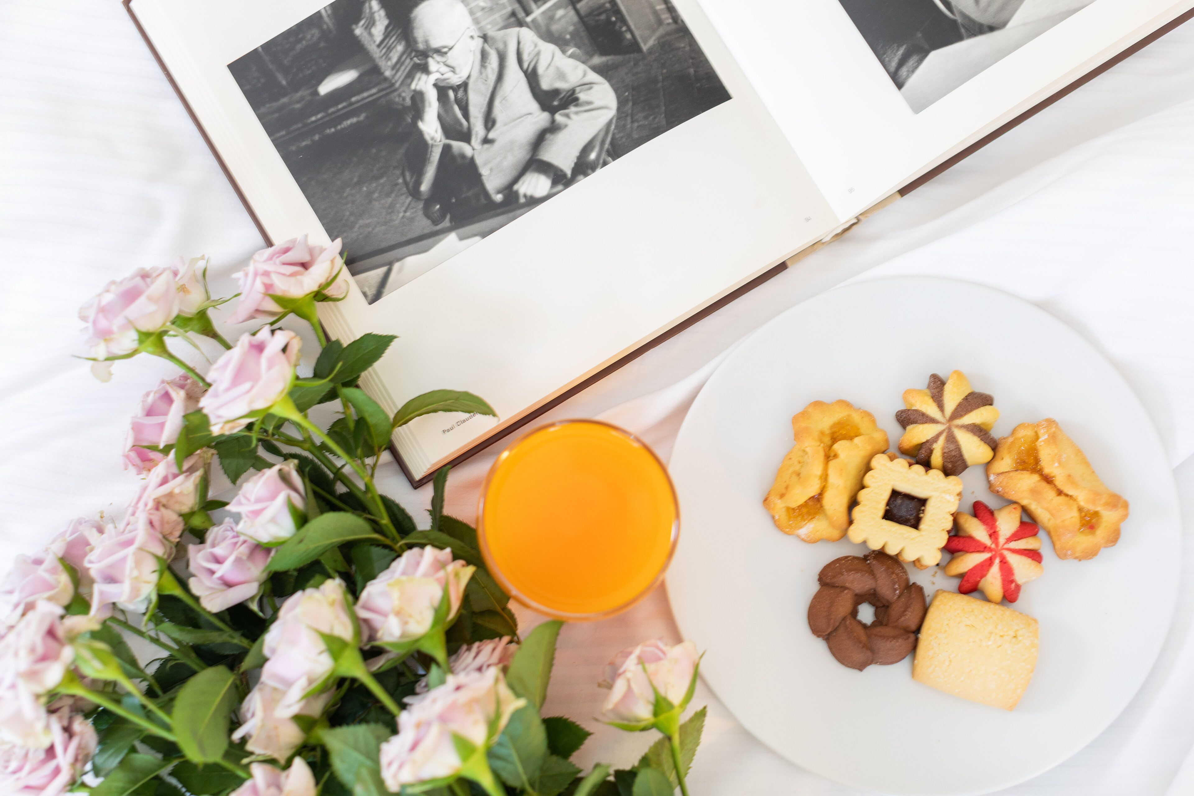 Un libro, un piatto con biscotti, un bicchiere di succo d'arancia e rose rosa su una superficie bianca.