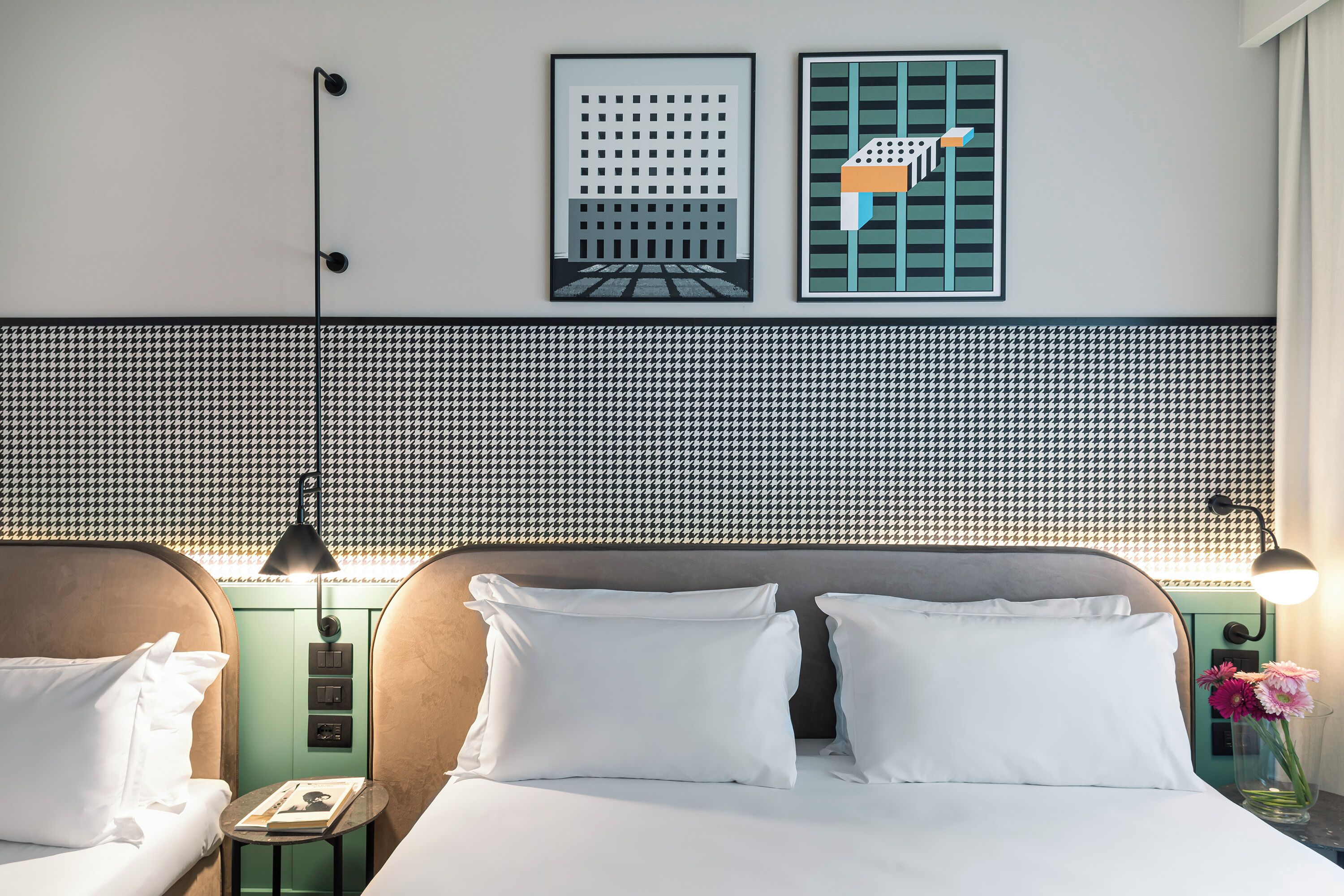 Camera d'albergo con una testiera a motivi a caccia, due letti con lenzuola bianche, un comodino con lampada e un punto luce a parete.