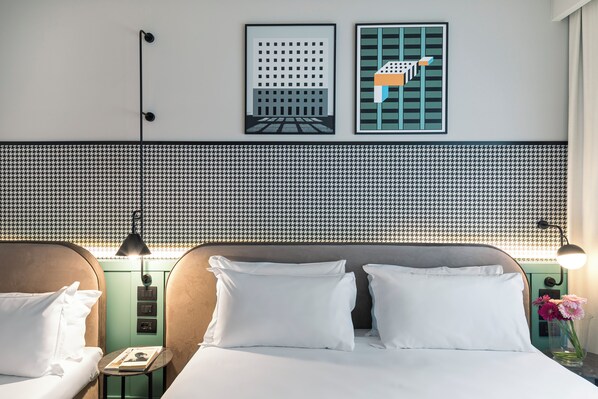A hotel room with a houndstooth-patterned headboard, two beds with white linens, a small bedside table with a lamp, and a wall-mounted light fixture.