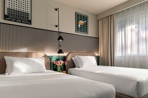 Een hotelkamer met twee bedden, een wand met een hondentandpatroon, een lijstwerk en een raam met dunne gordijnen.