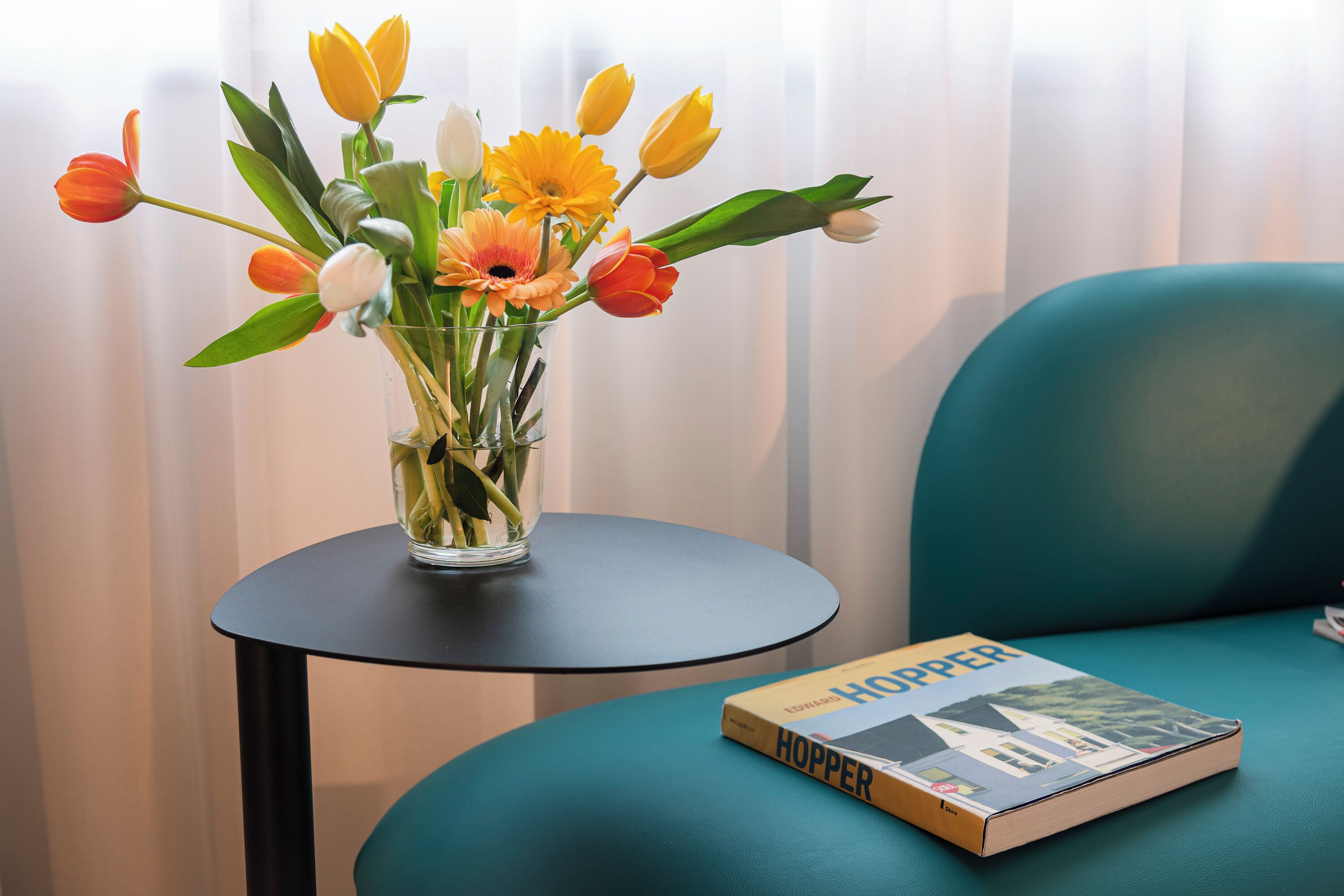 Um vaso com flores sobre uma pequena mesa ao lado de uma cadeira azul-esverdeada e um livro intitulado "Hopper".