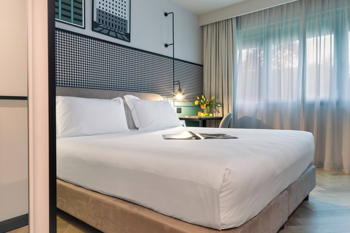 Ein modernes Hotelzimmer mit einem großen Bett, einem Schreibtisch mit Lampe und Blick ins Freie durch transparente Vorhänge.