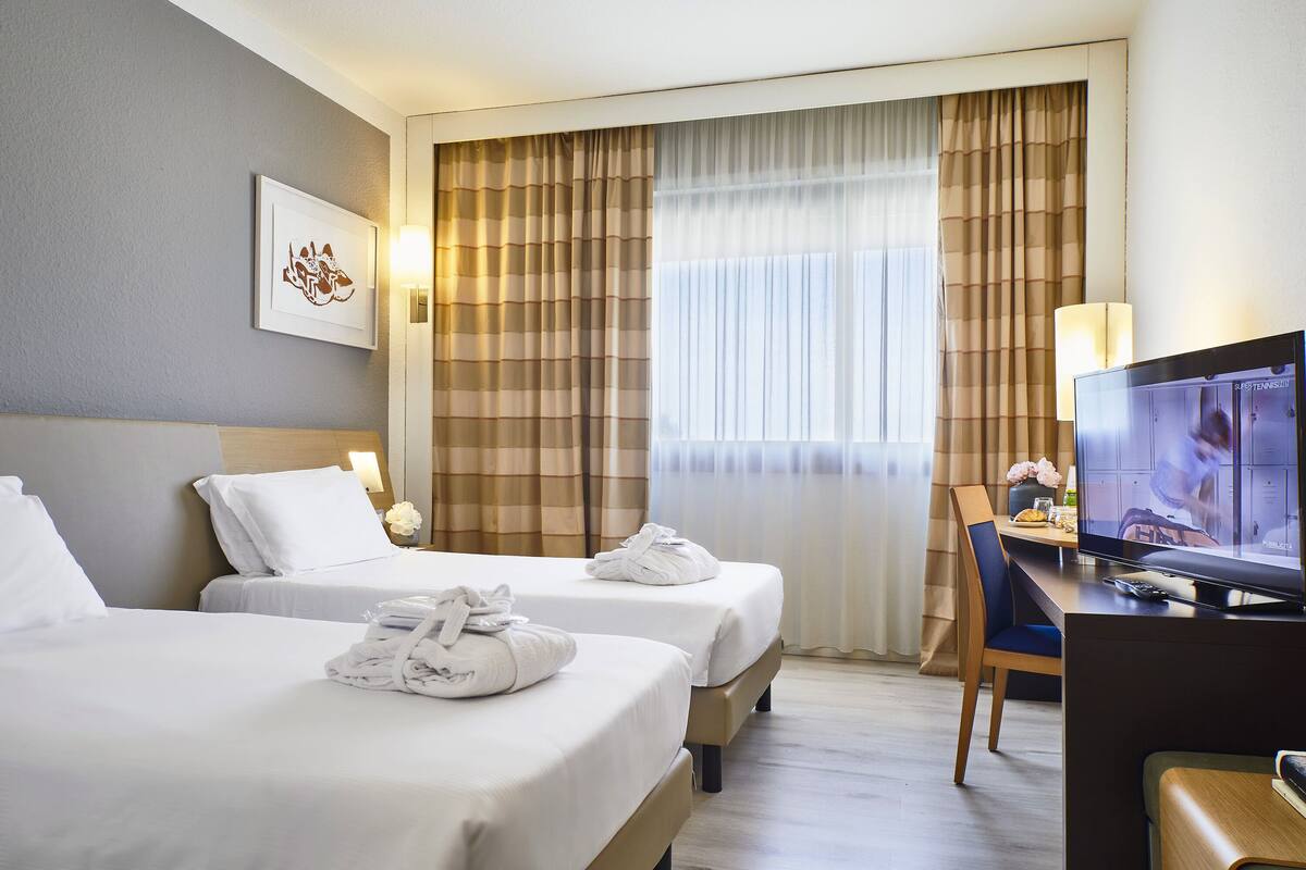 Ein Hotelzimmer mit zwei Betten, einem Schreibtisch mit Fernseher und einem Fenster mit Vorhängen.