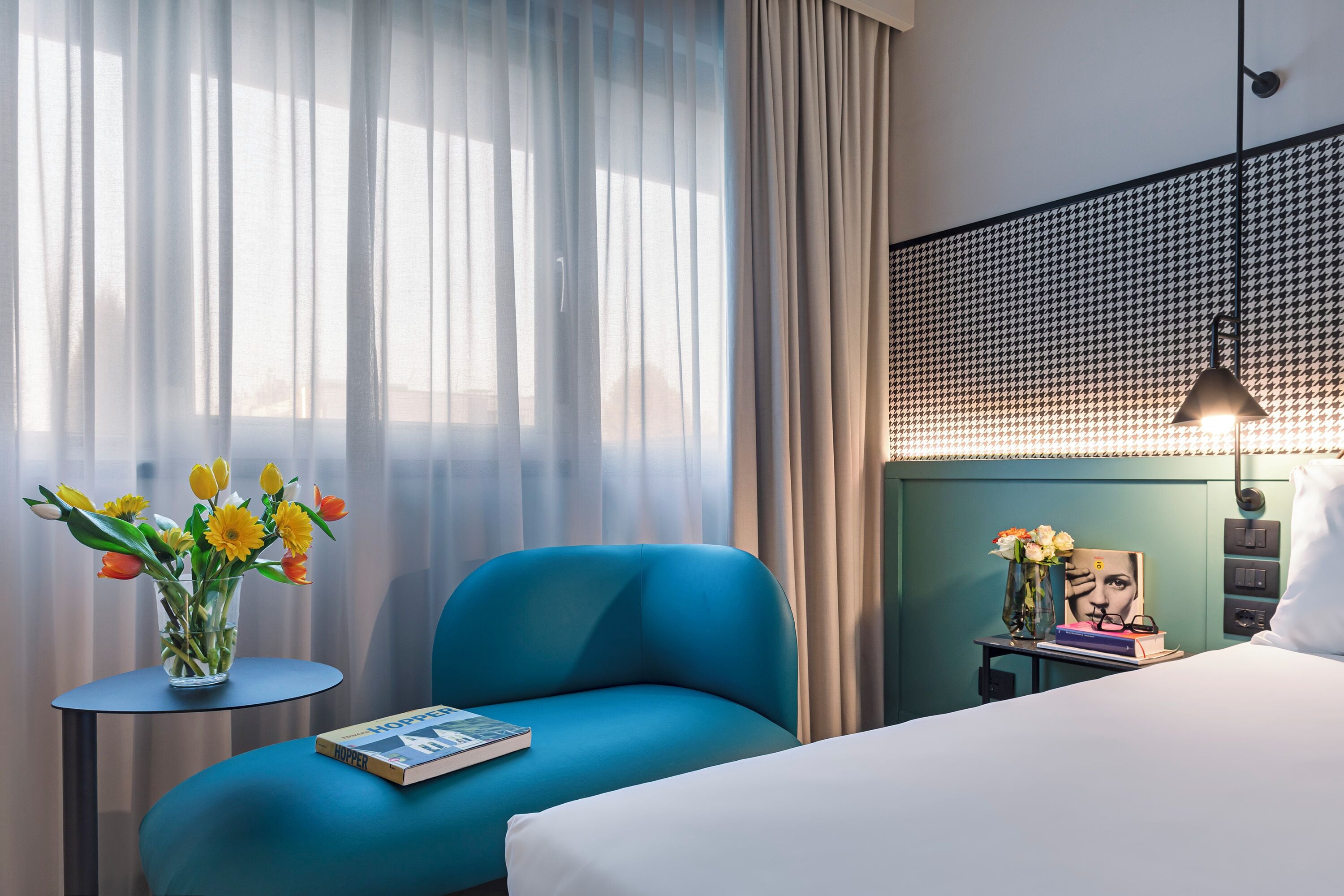 A hotel room with a bed, a blue armchair, a small table with a vase of flowers, and a wall with a houndstooth pattern.
