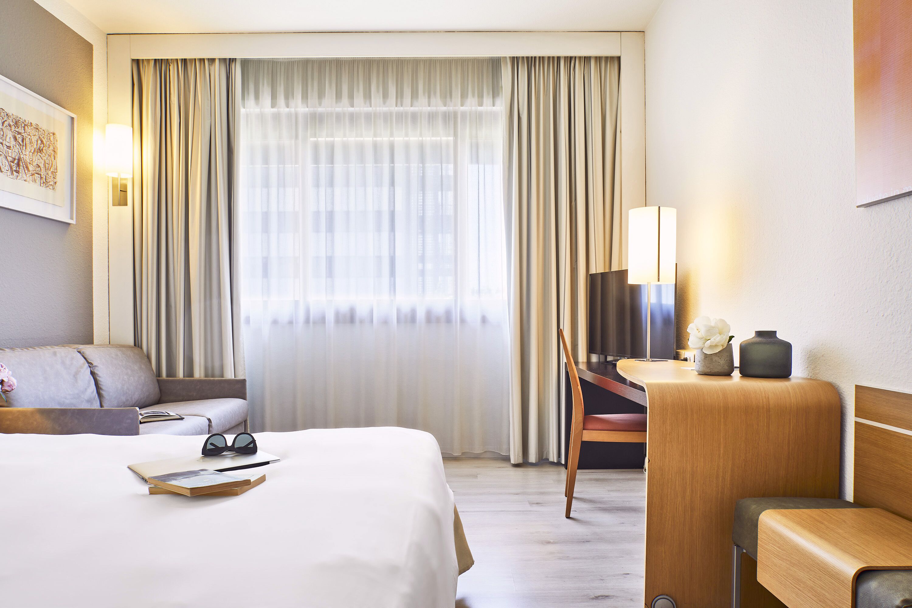 Een hotelkamer met een bed, een bureau, een stoel en een groot raam met gordijnen.