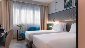 A hotel room with two beds, a desk, a chair, a small table with flowers, and a window with curtains. - Living Place Hotel (Bologna)