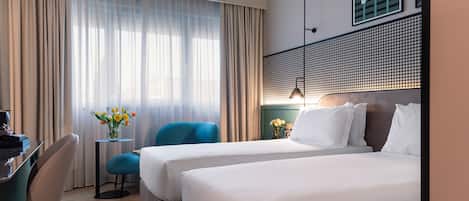 A hotel room with two beds, a desk, a chair, a small table with flowers, and a window with curtains.