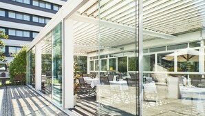 A modern outdoor dining area with glass walls, tables, and chairs, overlooking a cityscape. - Living Place Hotel (Bologna)