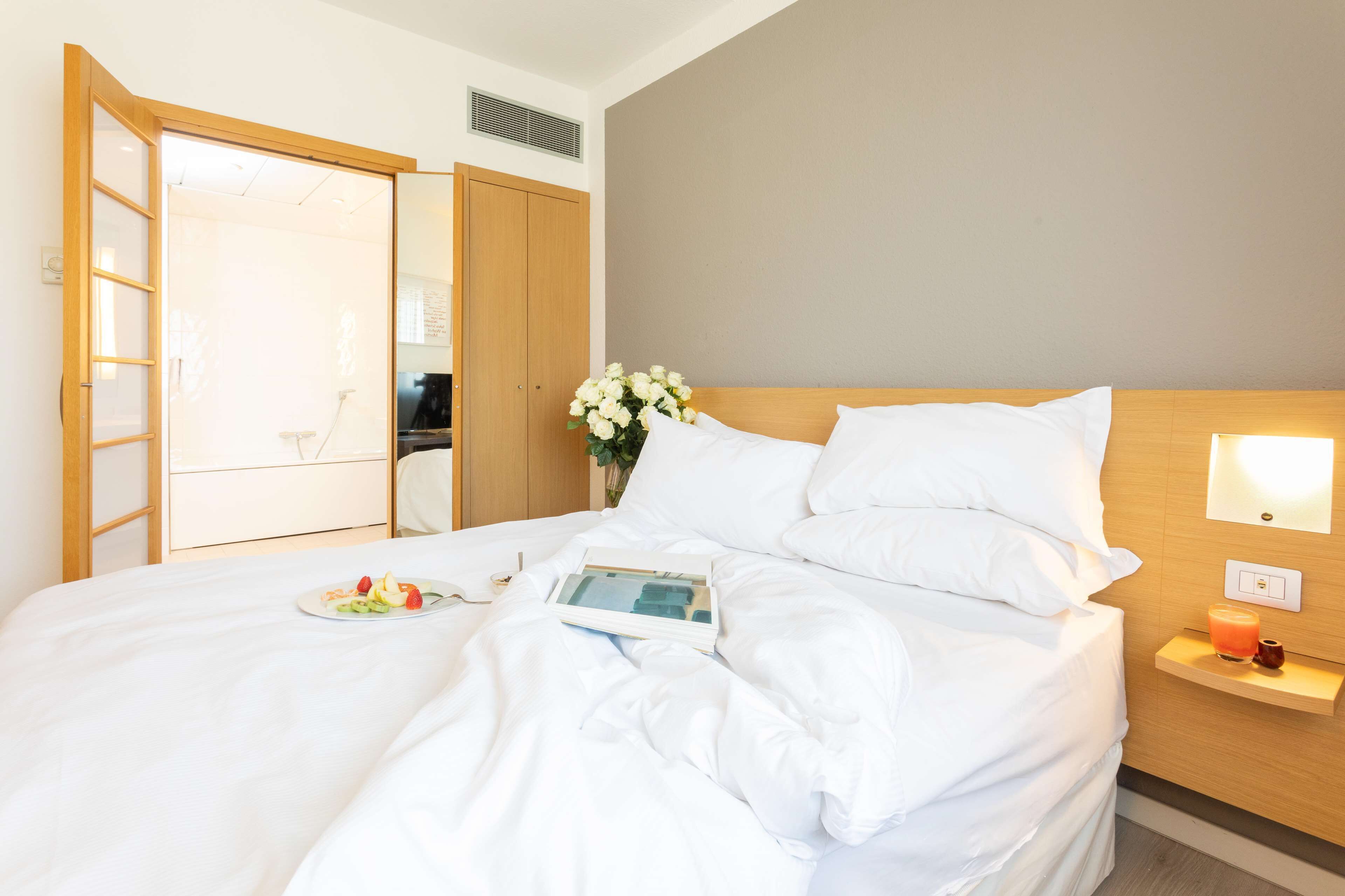 Una cama bien hecha con sábanas blancas, una mesita de noche con una vela y un vaso de zumo de naranja, y una lámpara de pared.