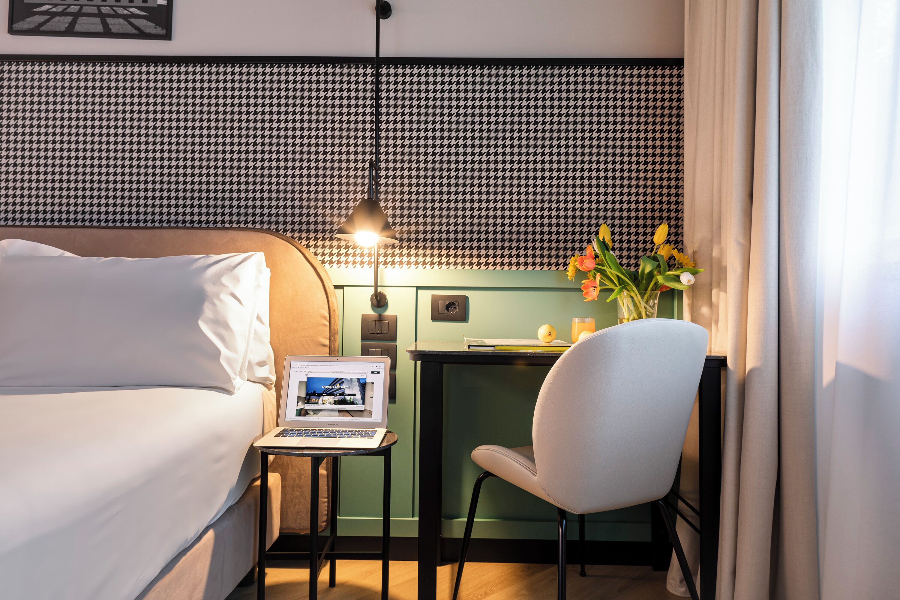 A hotel room with a bed, a desk with a laptop, a chair, a vase with flowers, and a patterned headboard.