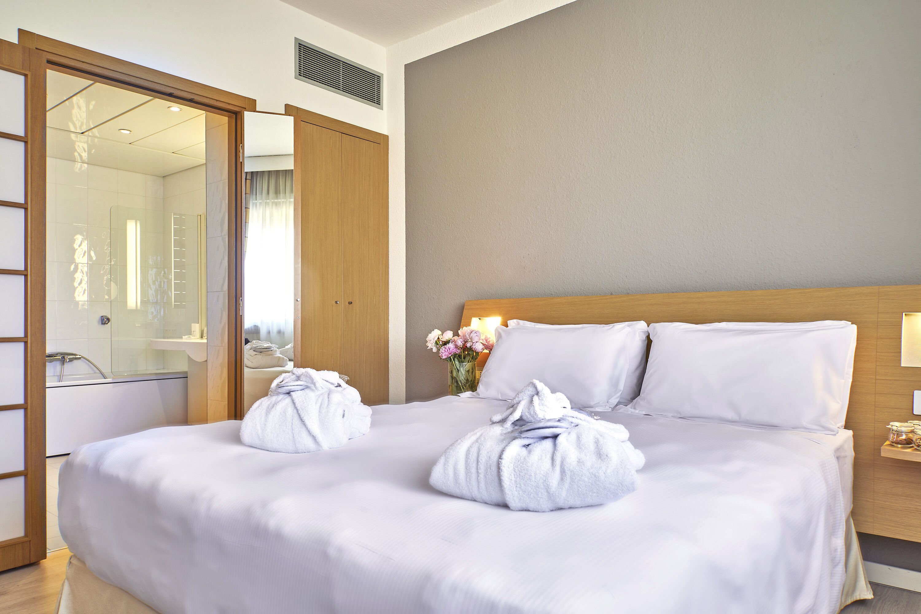 Habitación de hotel con una cama grande, cabecera de madera, mesita de noche con lámpara y baño visible a través de una puerta abierta.